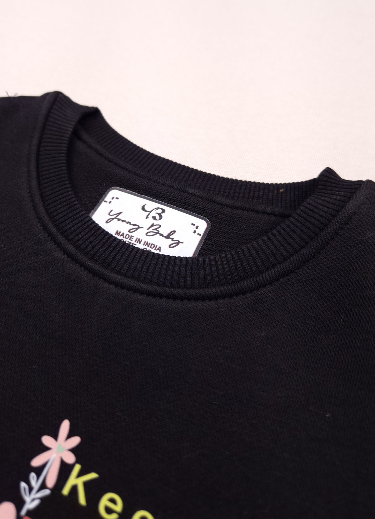 Close-up of a black crewneck sweatshirt showing the inner neck label which reads 'Young Baby' and 'MADE IN INDIA'. The bottom of the sweatshirt features a small graphic with a pink flower and the partial text 'Keep'.
